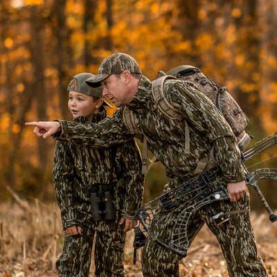 A man and boy wearing Mossy Oak Camo are in the woods bowhunting. The father is pointing to something in the distance and explaining it to his son.