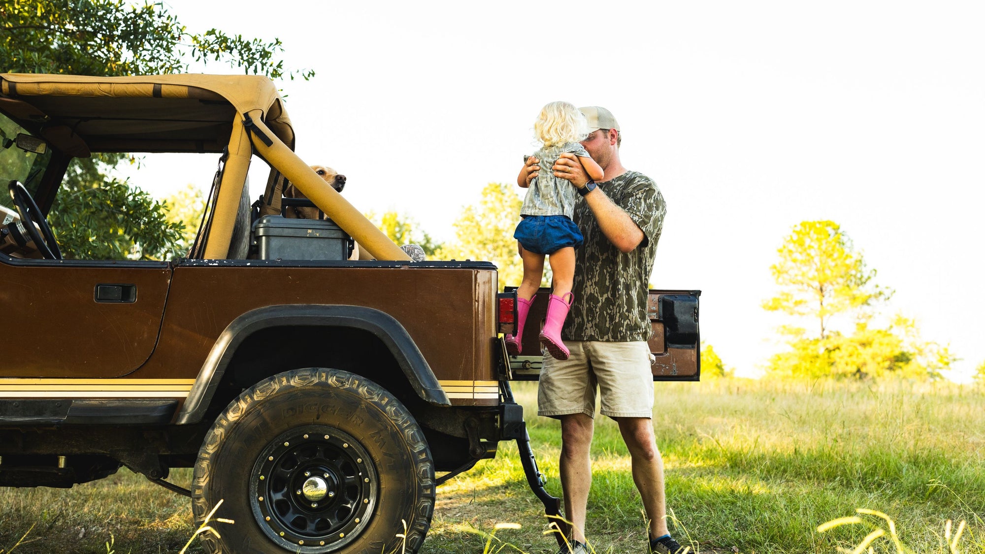 Man in camo shirt lifting child with blonde hair and pink boots into the back of a brown off-road vehicle outdoors