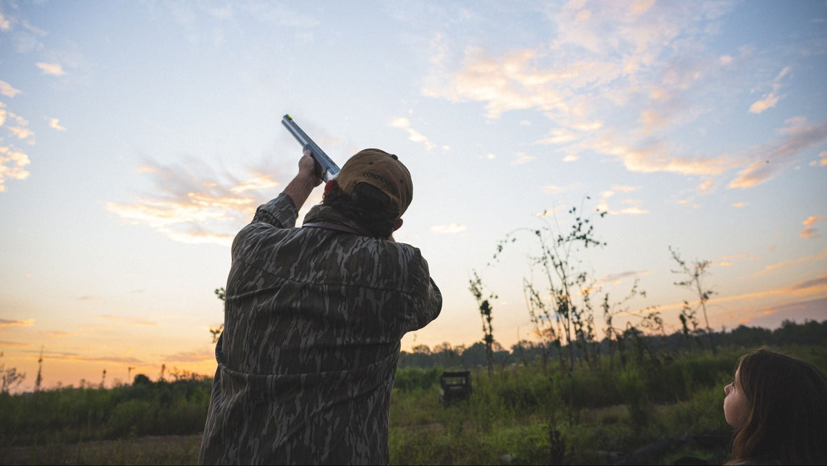 Hunter in camouflage aiming a shotgun at dusk while another person watches in a field
