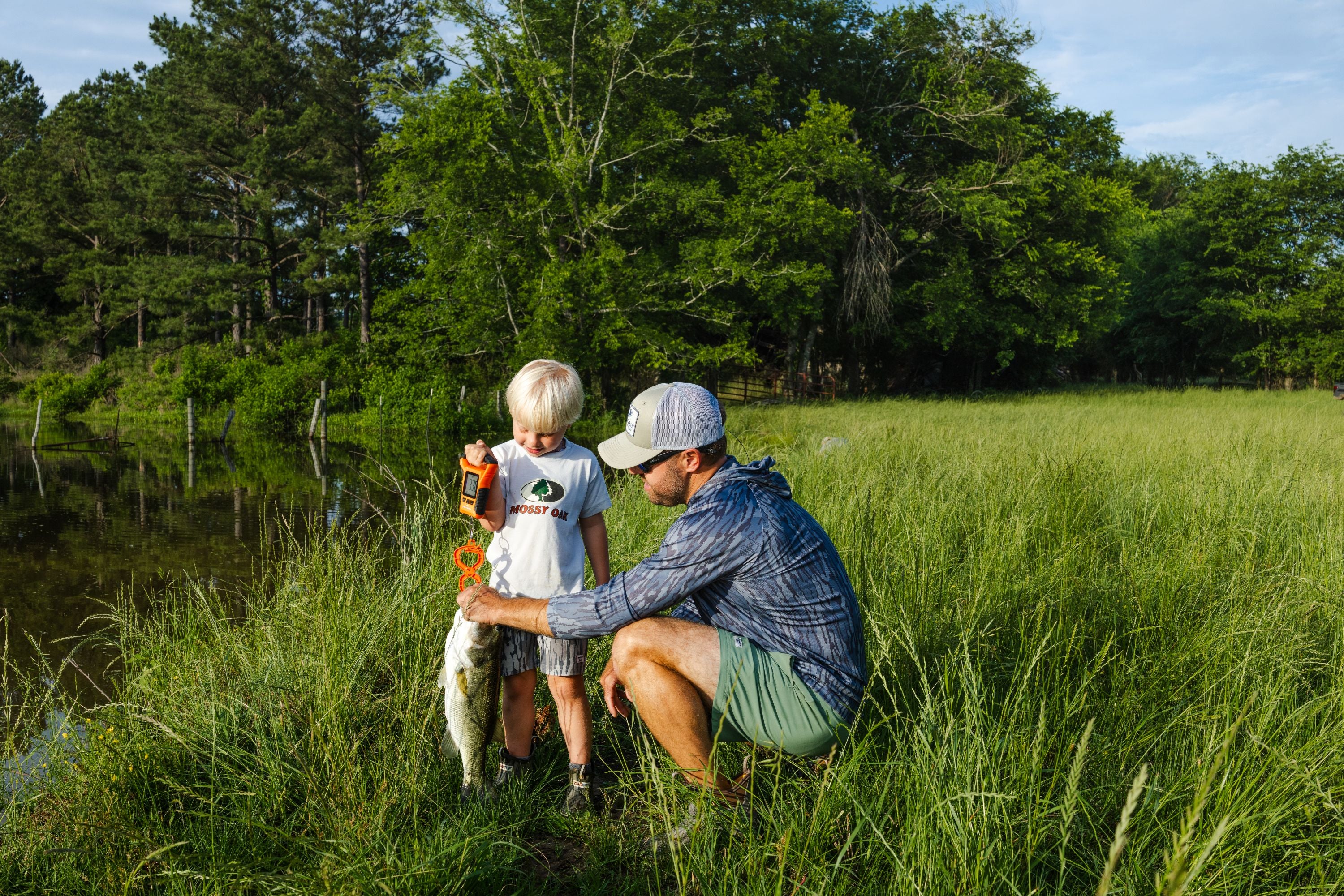 Man in casual outdoor clothing kneels beside young boy holding a large fish near a green pond and trees.