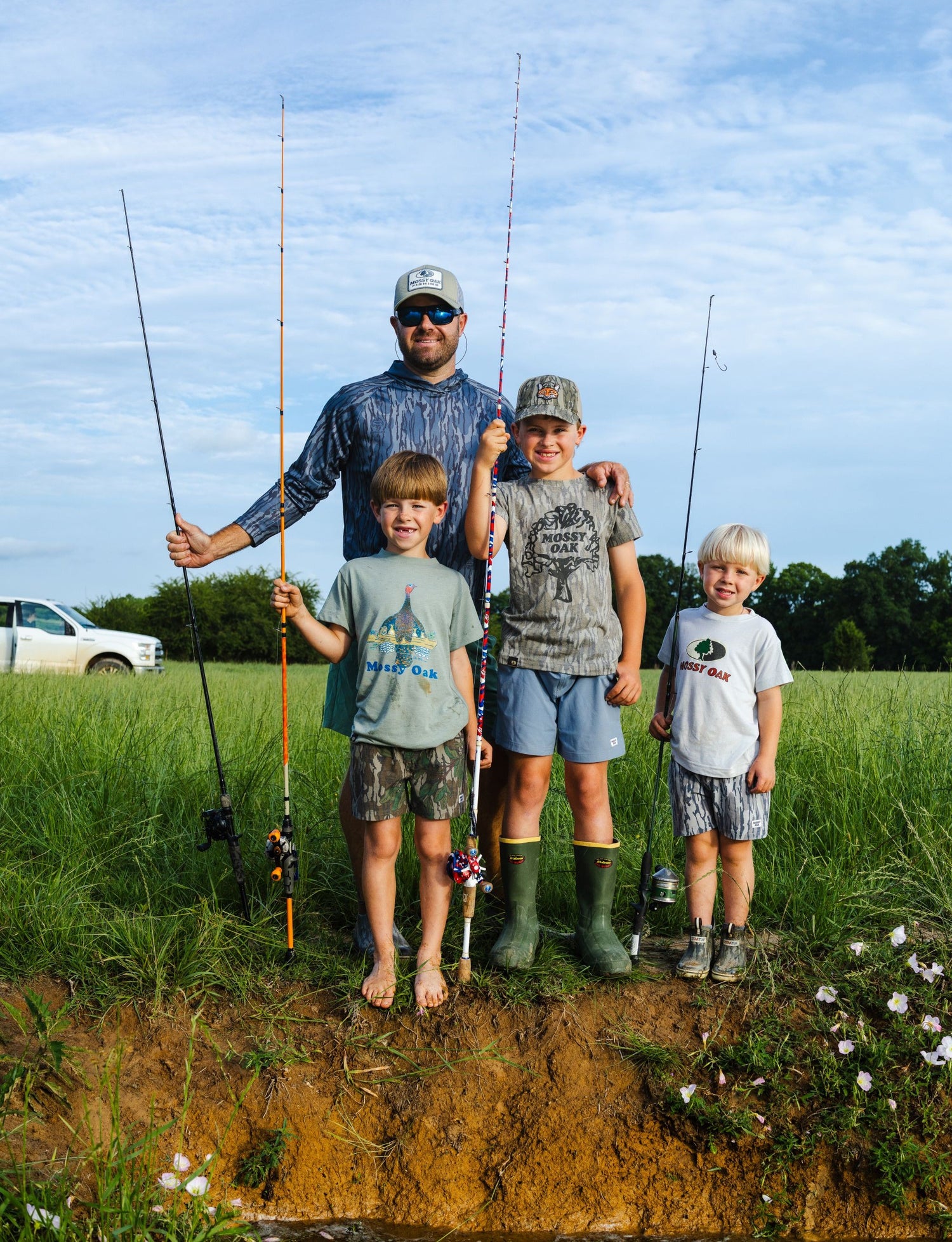 Man and three boys standing in grass holding fishing rods by a water edge on a clear day