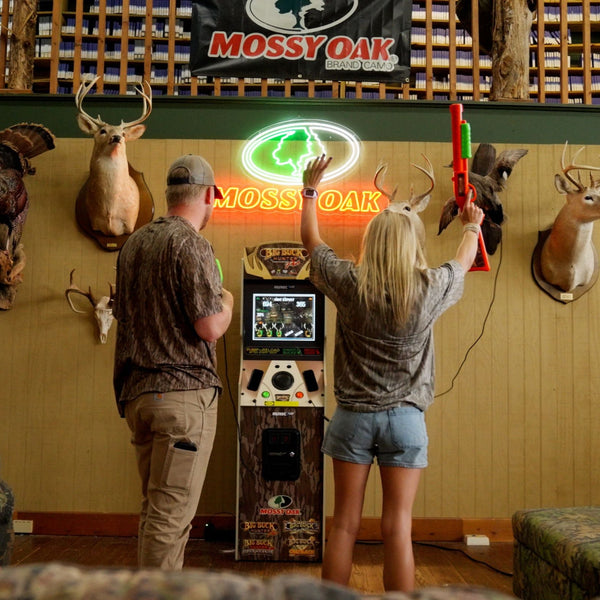 People playing a video game in a room with hunting-themed decor, including deer heads and a Mossy Oak sign.